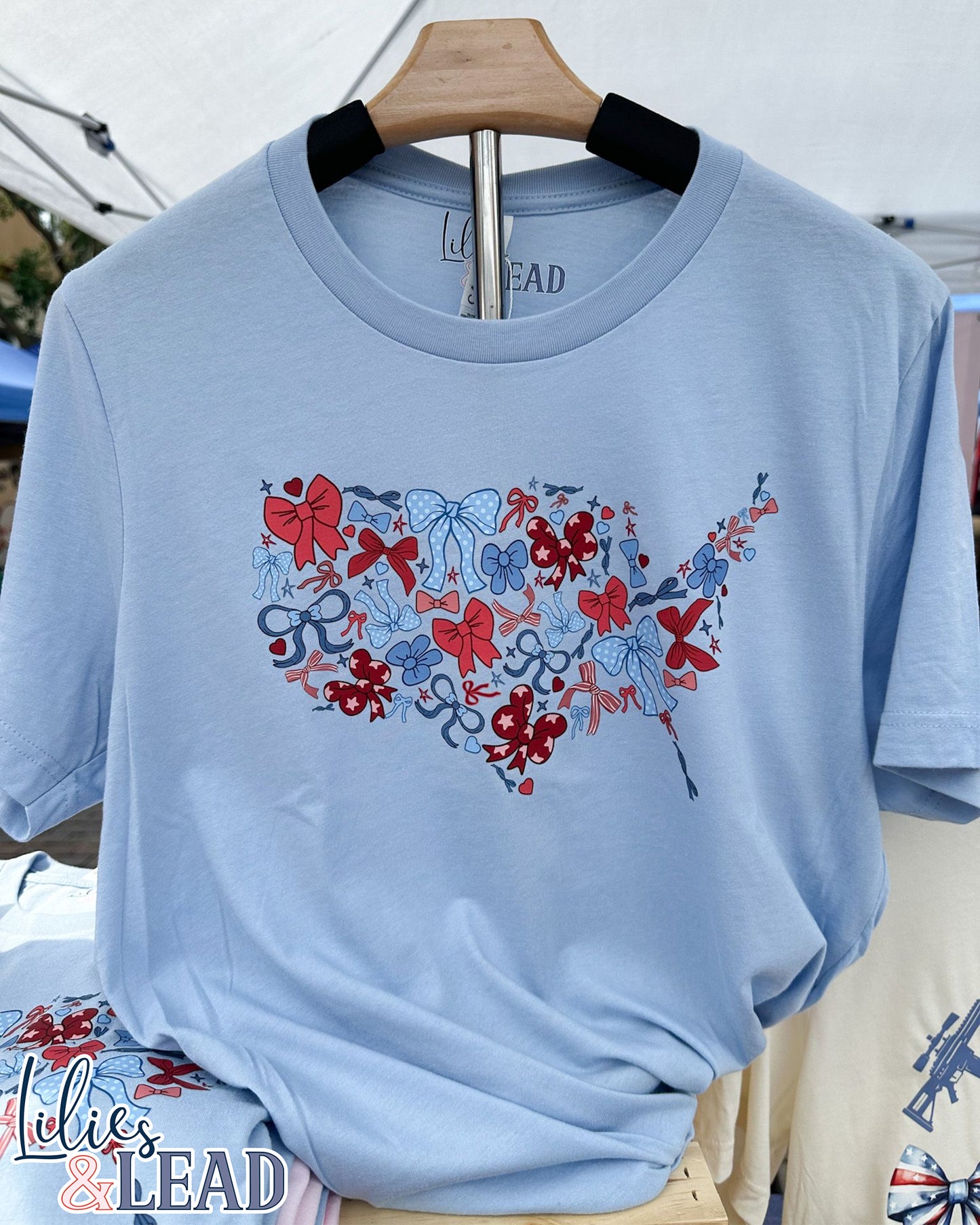 Light Blue Short sleeve Tee with USA Map design made from red, white and blue bows hanging on display.