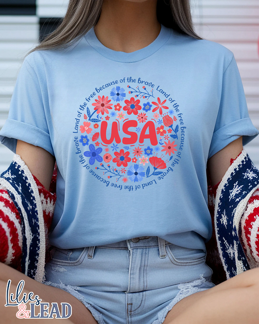 Light blue short sleeve tee shirt featuring "USA" in red surrounded by red white and blue floral encircled in the phrase "Land of the free because of the brave".