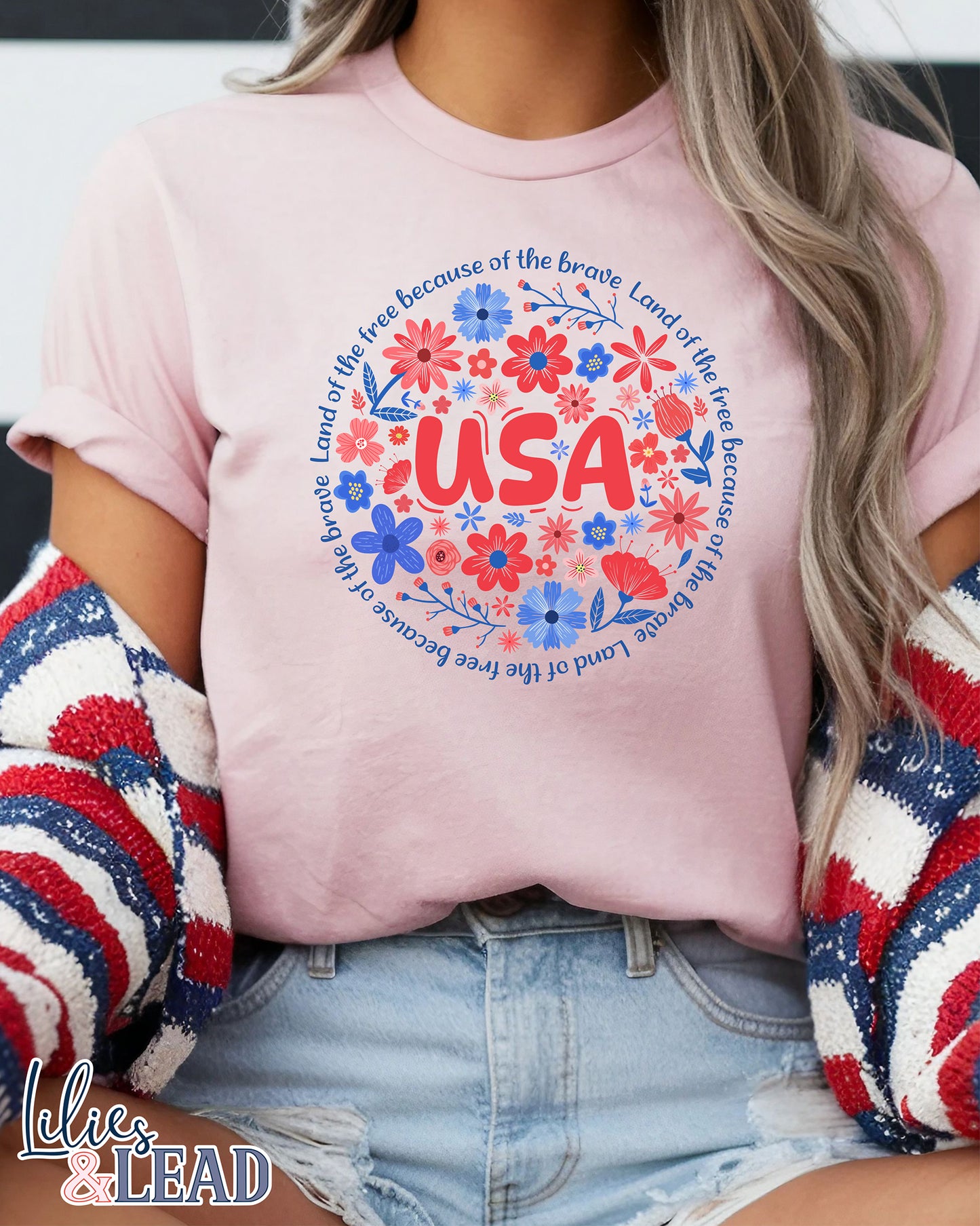 Light pink short sleeve tee shirt featuring "USA" in red surrounded by red white and blue floral encircled in the phrase "Land of the free because of the brave".