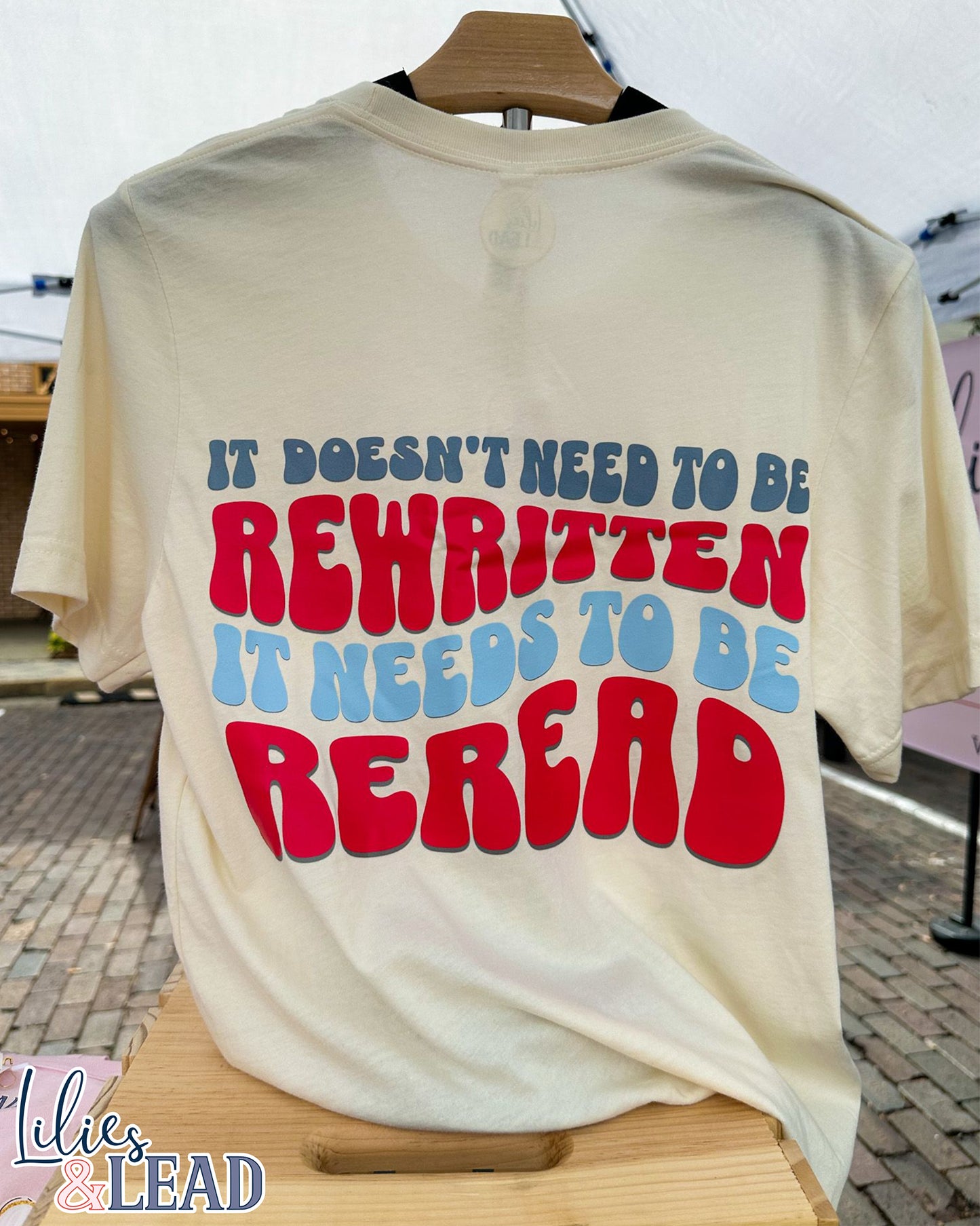 Short Sleeve Natural colored tee with the phrase It Doesn't need to be rewritten, it needs to be reread phrase in a retro font with Red, White, and blue