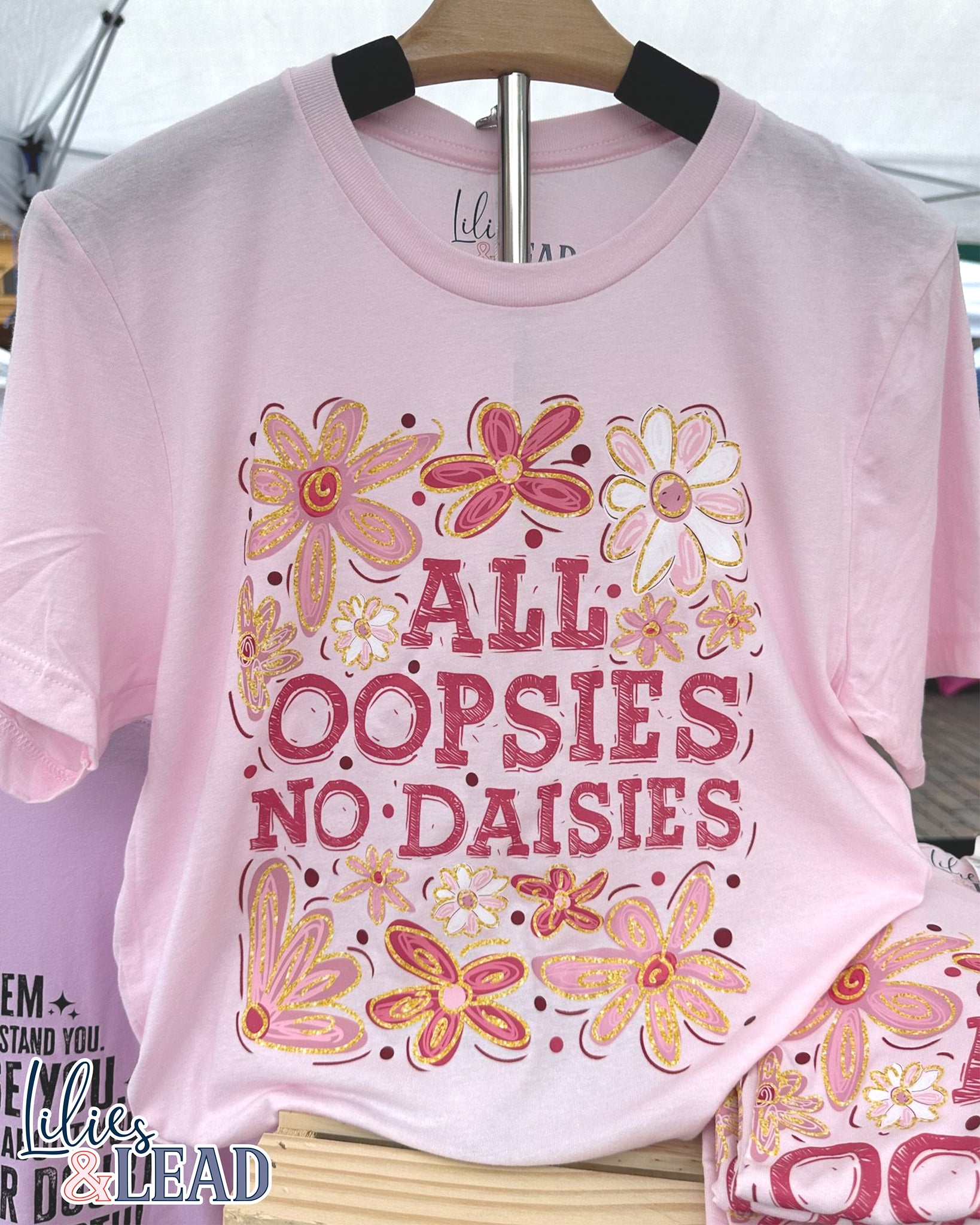 Light pink tee with saying All Oopsies No Daisies surrounded by florals in pink and white hanging on display.