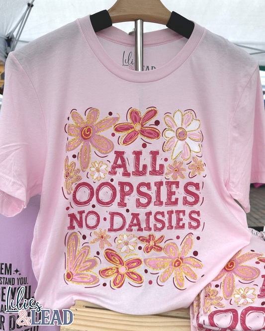Light pink tee with saying All Oopsies No Daisies surrounded by florals in pink and white hanging on display.