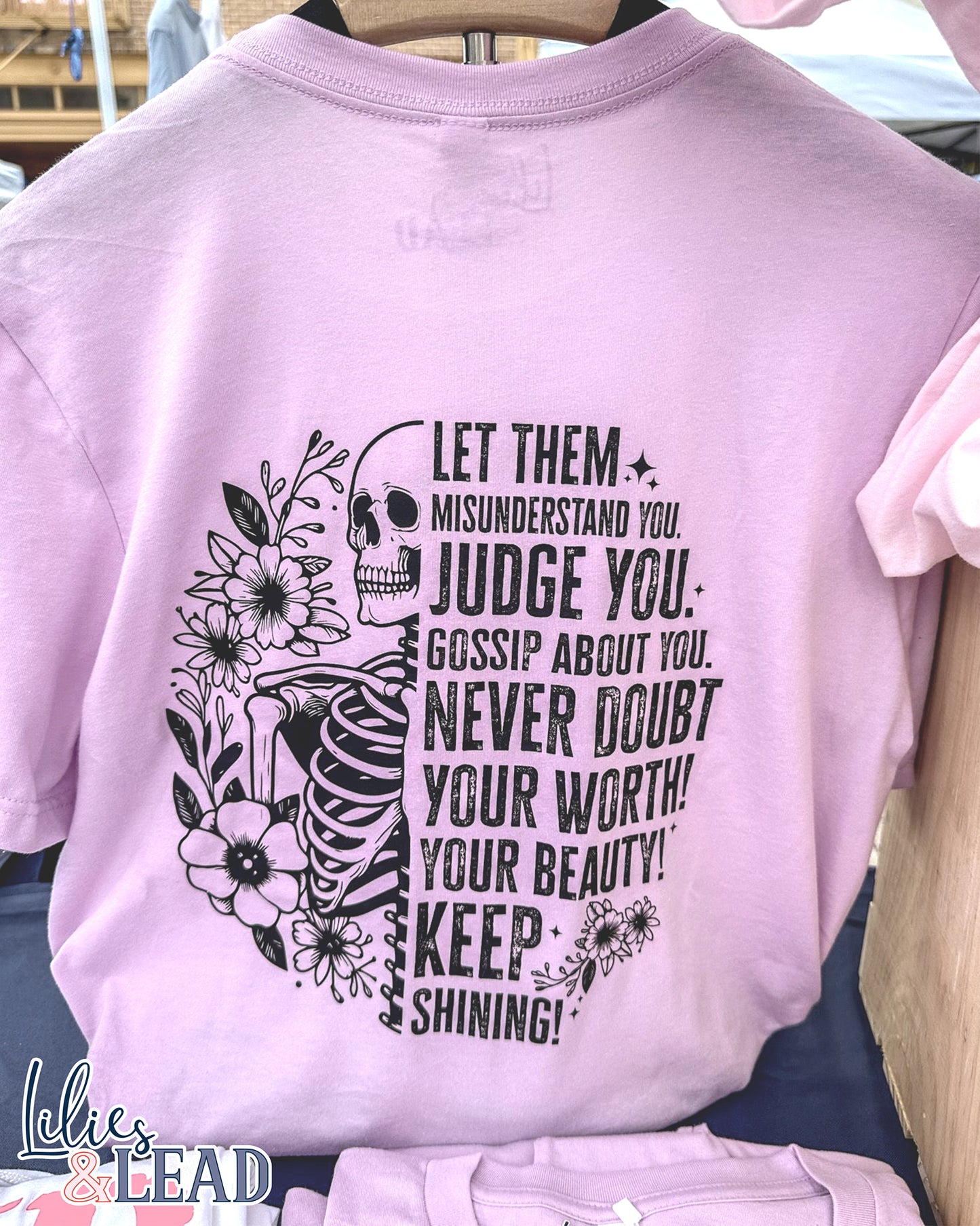 Lilac short sleeve tee with black skeleton and florals and the saying "Let them misunderstand you, judge you, gossip about you. Never doubt your worth! Your Beauty! Keep Shining!" Hanging on display.
