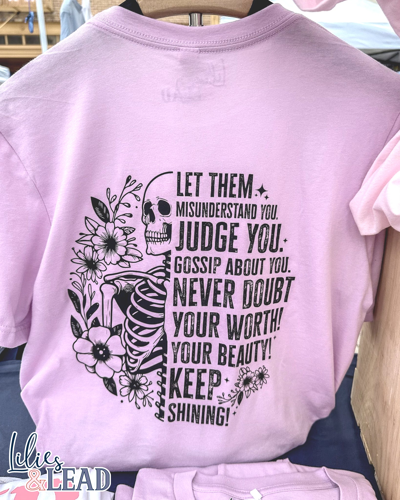 Lilac short sleeve tee with black skeleton and florals and the saying "Let them misunderstand you, judge you, gossip about you. Never doubt your worth! Your Beauty! Keep Shining!" Hanging on display.
