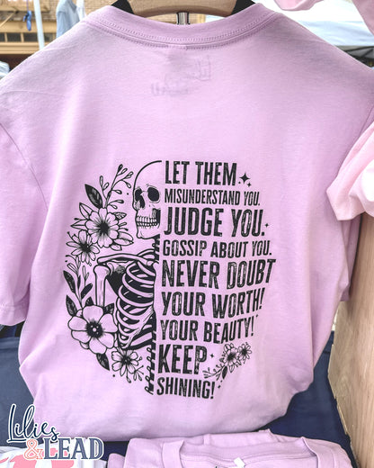 Lilac short sleeve tee with black skeleton and florals and the saying "Let them misunderstand you, judge you, gossip about you. Never doubt your worth! Your Beauty! Keep Shining!" Hanging on display.