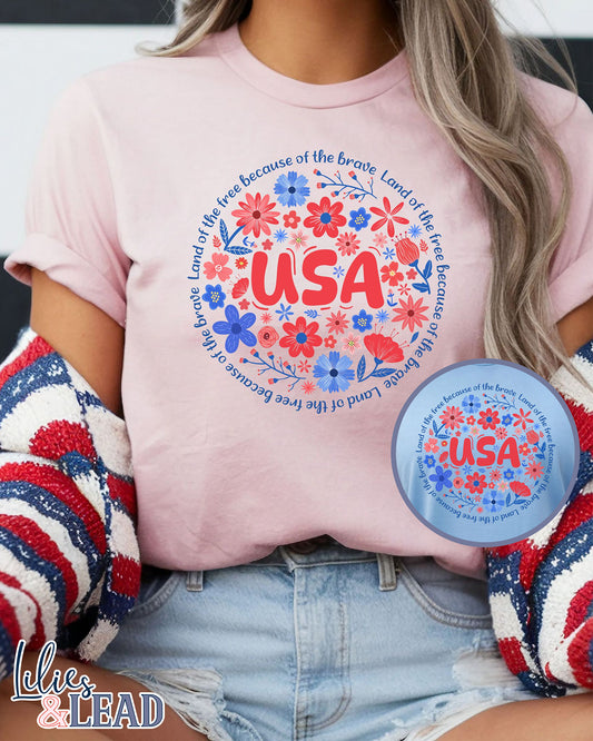 Light pink short sleeve tee shirt featuring "USA" in red surrounded by red white and blue floral encircled in the phrase "Land of the free because of the brave".